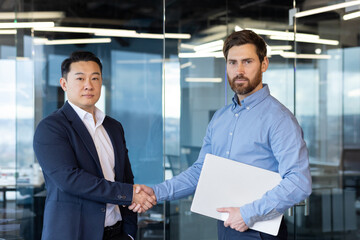 Two diverse businessmen shaking hands in a bright office setting, illustrating partnership and corporate agreement between colleagues.