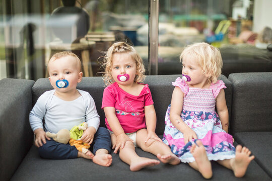 Three young kids sitting on couch all with pacifiers in their mouths