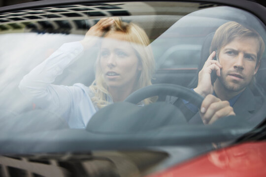 Couple in business attire driving electric sports car and talking on the phone with worried look on their faces. Munich, Bavaria, Germany