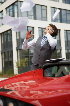Man in business attire standing next to electric sports car with papers flying around. Munich, Bavaria, Germany
