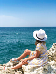 Little girl sitting on rocky shore while looking at sea during summer vacation with family on seaside