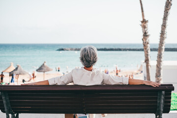 Rear view of relaxed senior woman sitting outdoor in a bench looking at the horizon over water. Beach and sea on background