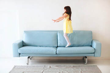 Little caucasian cute girl in yellow dress playing and jumping on blue sofa in living room at home
