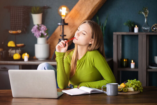 Young pensive girl sitting at table at home with laptop, making notes, staring thoughtfully into air
