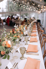 A beautiful set table is decorated in peach tones with flowers on the table and golden glasses, plates with napkins. Festive set table for guests without people with garland in the background