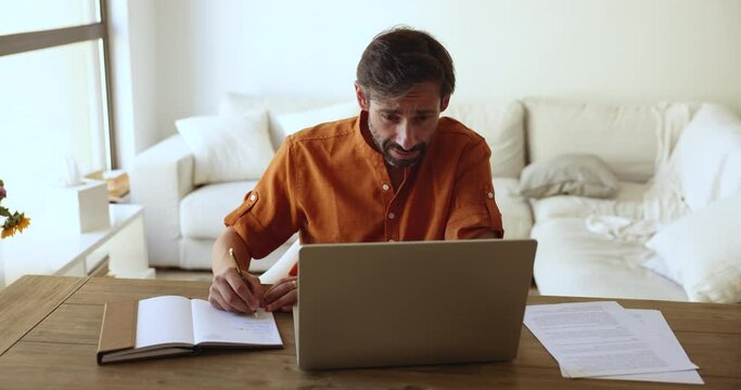 Serious focused middle-aged Portuguese man writing important information in personal organizer, take notes or to-do list in paper planner, make task sit at desk staring at computer screen. Workflow