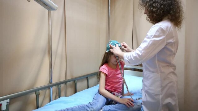 doctor in lab coat puts cap for encephalography on girl head