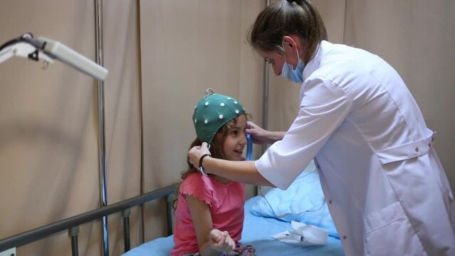 Female doctor puts special cap for encephalography on girl head.