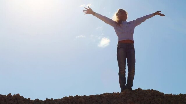 Girl stands on stone pile and waves by hands during sunny day