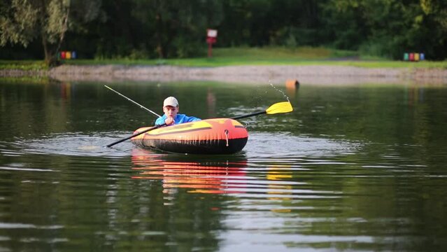 Boy paddles in inflatable boat with fishing rod in pond at summer
