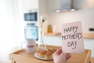 Mother's day still life with mom cup, a sweet pastry and a hand holding a greeting card, in airy indoor background.