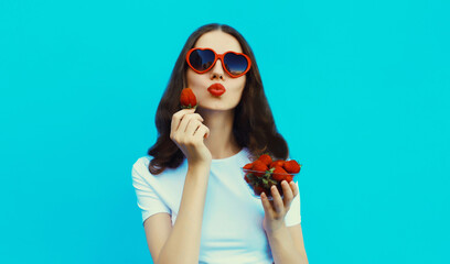 Portrait of beautiful caucasian young woman with handful of fresh strawberries blowing her lips wearing red heart shaped sunglasses on blue studio background - obrazy, fototapety, plakaty