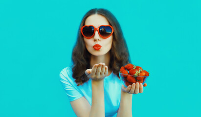 Portrait of beautiful caucasian young woman with handful of fresh strawberries blowing her lips wearing red heart shaped sunglasses on blue studio background