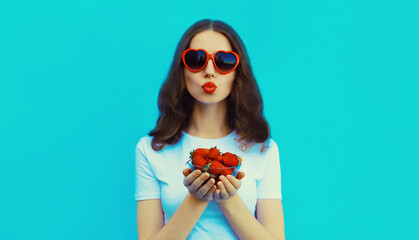 Portrait of beautiful caucasian young woman with handful of fresh strawberries blowing her lips wearing red heart shaped sunglasses on blue studio background - obrazy, fototapety, plakaty