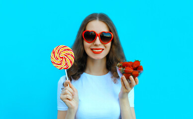 Portrait of beautiful caucasian young woman with handful of fresh strawberries holding sweet lollipop wearing red heart shaped sunglasses on blue studio background - obrazy, fototapety, plakaty