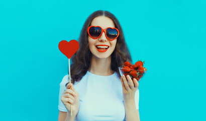 Portrait of beautiful caucasian young woman with handful of fresh strawberries holding sweet lollipop wearing red heart shaped sunglasses on blue studio background