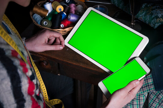 Over The Shoulder View Of A Woman Tailor Sewing With Help Of A Tablet And Smart Phone With Green Screen On Them