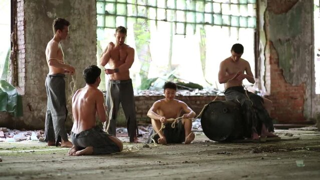 Five men in ragged pants with rope in abandoned building