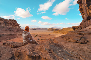 Young woman sitting on rocky ground in desert landscape, small vehicle far distance. View from behind. Wadi Rum, Jordan