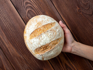 Close-up of hand holding a round whole loaf of wheat bread. Brown wooden background, Top view, flat lay. Bakery concept.