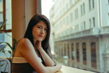 young Latin woman looks at the camera satisfied, sitting in a café with a view of the city. copy space