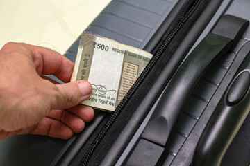 Closeup view of hand holding five hundred rupees banknotes and suitcase.