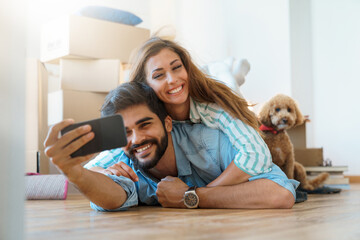 Affectionate young couple using mobile phone taking self portrait photos in new apartment while...