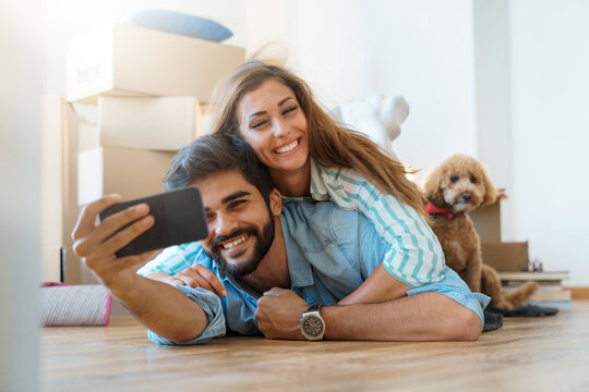Affectionate young couple using mobile phone taking self portrait photos in new apartment while lying on floor with dog.