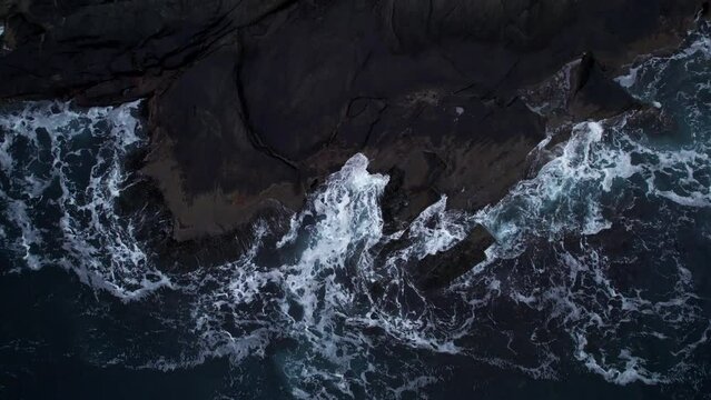 Top-down aerial shot of cold arctic ocean waves hitting rocky shoreline, zoom in
