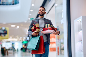 Happy man carrying wrapped presents and shopping bags while walking through mall.