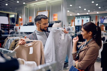 Happy man asking advice from his girlfriend while buying clothes in shopping mall. - obrazy, fototapety, plakaty