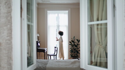 Domestic girl open curtains at morning indoors. African woman enjoying espresso