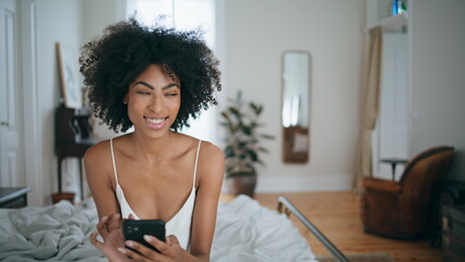 Smiling woman messaging smartphone at bedroom. Curly laughing lady sitting bed