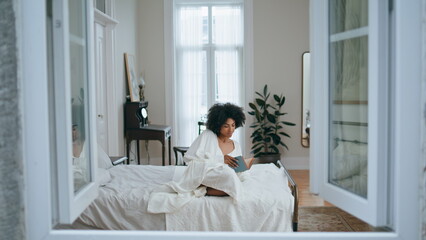 Carefree woman flipping book pages indoor. African lady reading novel laying bed