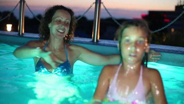 Woman and girl dance in blue pure pool on terrace in evening