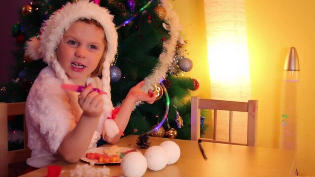 Little girl in santa cap paints on cookies near christmas tree