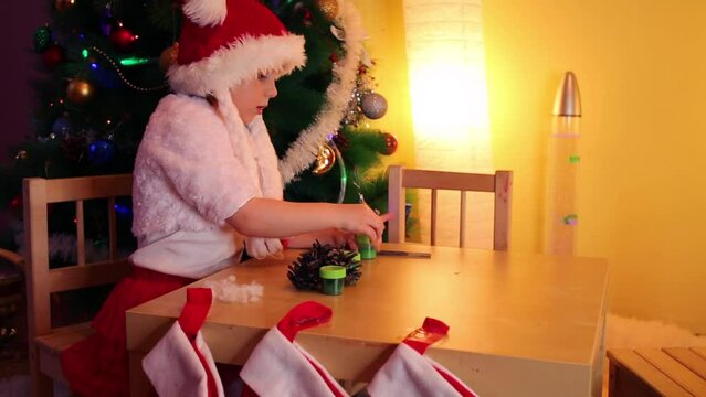 Little girl in santa cap sit at table near christmas tree and plays