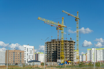 Fototapeta premium Silhouettes of working construction cranes against the background of an unfinished residential building. Multi-storey luxury apartment building on a construction site