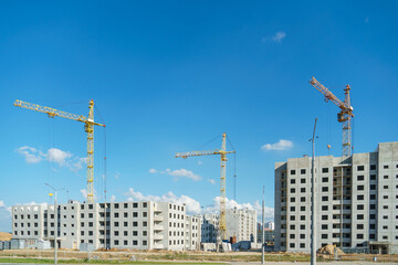 Fototapeta premium Silhouettes of working construction cranes against the background of an unfinished residential building. Multi-storey luxury apartment building on a construction site