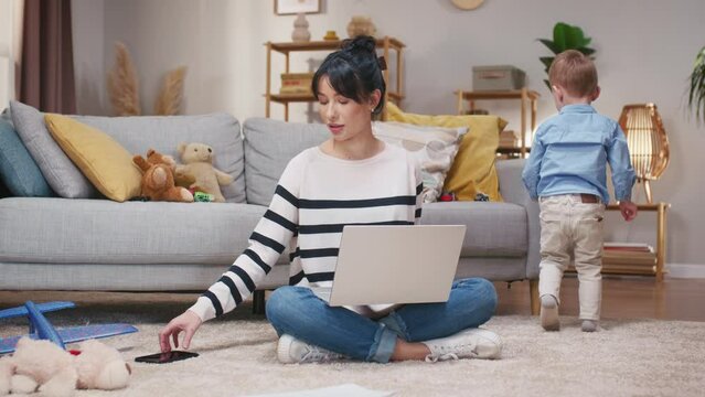 Woman freelancing while on maternity leave. Mother working with laptop and taking care of son. LIttle boy playing behind. Mother sitting at carpet floor and using laptop. Boy getting boring near mom.