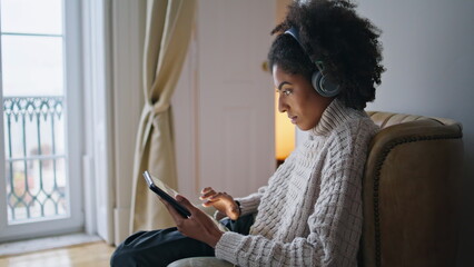 Focused girl scrolling pad comfy home. African headphones lady listening music