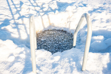 Bright winter day with a frosted ladder leading into a natural ice swimming hole on a snow-covered...