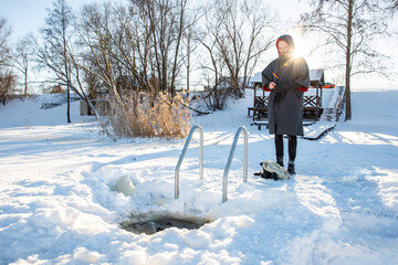 Man in winter gear standing by a swimming hole cut in the snow, with a serene winter landscape...
