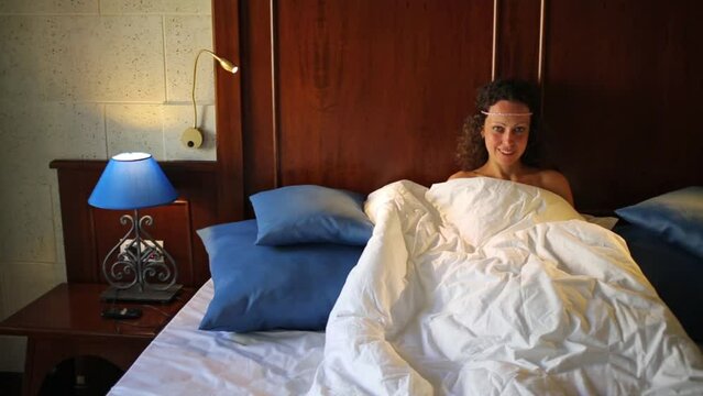 Pretty woman sits under white blanket on wooden double bed