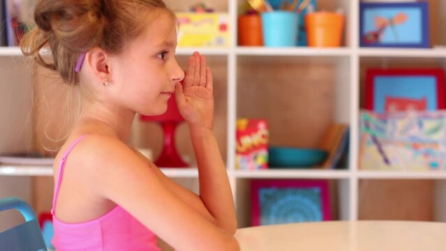 Little girl sits at table and raises hand in play room with shelves