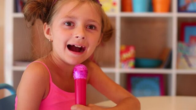Little girl singing into toy microphone sitting at table in room