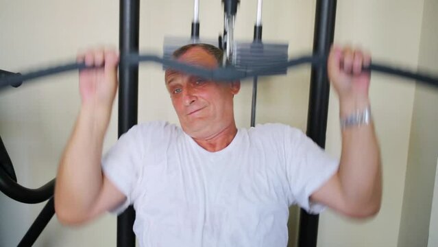 Elderly man training on exerciser for arms in fitness center