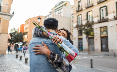 couple hugs on the street, she with a bouquet of roses that can be seen on the boyfriend's back...