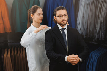 Beautiful woman seller smiling and help with choice suit to modern young handsome businessman in shop of classic clothes for men