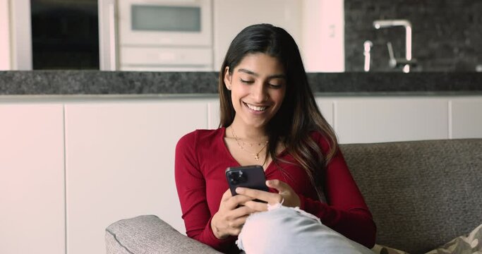Smiling Indian woman hold smart phone resting seated on sofa in living room. Young generation person using modern wireless technology for fun, leisure, remote communication, electronic services usage